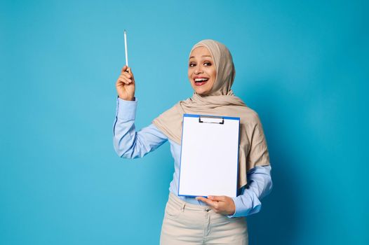 Smiling Arab woman in hijab holding a clipboard with a white blank sheet of paper and points a pen on blue background with space for text