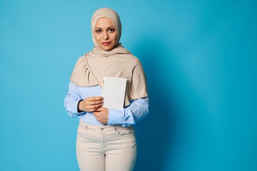 Serious and serene Arab Muslim woman with a white book in her hands. Education concepts on blue background with copy space