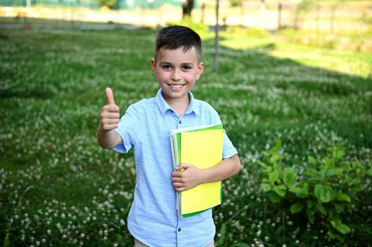 Adorable schoolboy holding a yellow workbook and showing thumb up to camera standing on green grass background. Happy elementary student coming back to school
