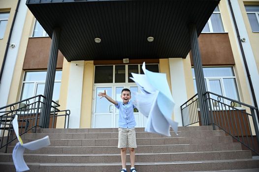 Elementary student, boy rejoices at the end of lessons and school, throws workbooks up, laughing and smiling happily while standing on the stairs against the background of the school institution