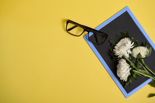 Flat lay of delicate beautiful bouquet of asters autumn flowers and eyeglasses on a blank empty chalkboard with space for text isolated on yellow background
