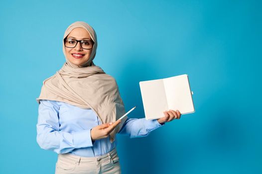 Smiling Muslim woman in hijab holds blank book and points at it with pen isolated on blue background with space for text