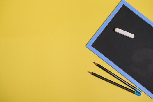Flat lay composition with chalk on a clean blank blackboard with copy space and two color pencils, isolated on yellow background with space for text