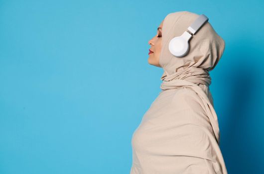 Side view of Arab Muslim woman in hijab and headphones posing over blue background with copy space