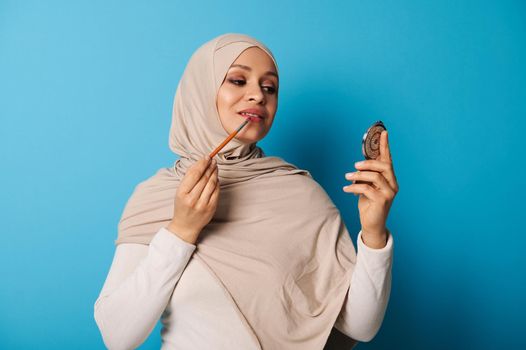 Young Arab woman in beige hijab uses a makeup brush to apply lipstick to her lips and looks at her reflection in a small cosmetic mirror. Isolated portrait on blue background with space for text