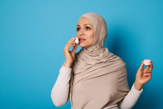 Charming woman in strict formal outfit and head scarf applying a lipstick, standing isolated over blue background with space for text