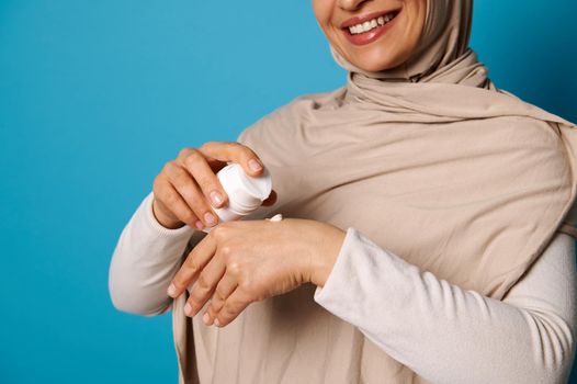 Closeup of smiling woman in hijab squeezing cream on hand, isolated on blue background with space for text