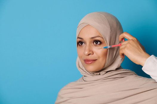 Attractive young Muslim woman with perfect makeup and clean healthy skin, posing with a syringe near her face over blue background. Space for text