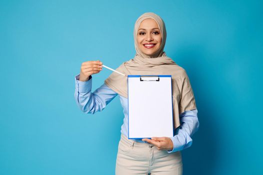 Smiling Arab woman with covered head in hijab pointing a pen on a blank surface on a white paper sheet in clipboard