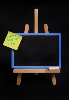 Close-up of table easel with chalk and blackboard with empty blank space for text and pasted note paper with inscription teachers day isolated on black background with copy space