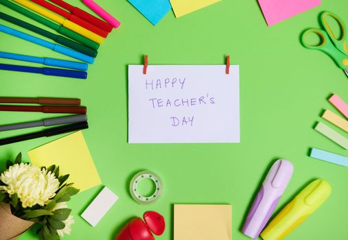 High angle view of colorful assortment of stationery office supplies and school accessories arranged in a circle, with white paper in the center with lettering Happy Teacher's Day.