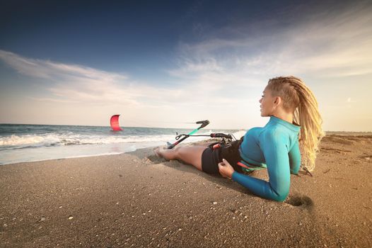 Attractive caucasian woman with dreadlocks on her head in a wetsuit lies on a sandy beach and holds her kite. Water sports. Kite surfer on vacation. Copy space.