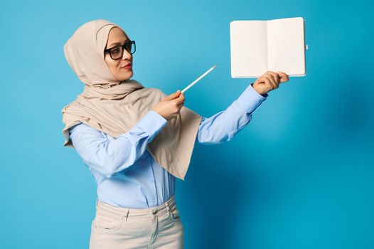 Muslim woman in hijab holds blank book and points at it with pen isolated on blue background with copy space