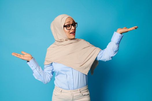 Arab woman in hijab posing with outstretched arms and palms up, presenting on blue background with space for text