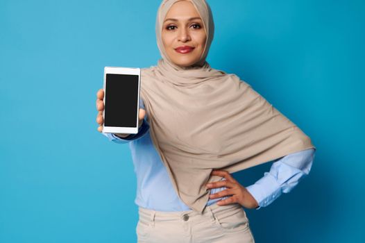 Soft focus on mobile phone in Arab Muslim woman's hands. Young woman in hijab holding a smartphone and posing over blue background with space for text