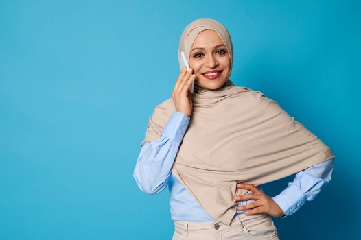 Isolated portrait of young Muslim woman in beige hijab talking on mobile phone. Confident portrait of a successful Arab woman on blue background with copy space