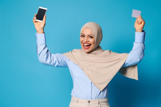 Isolated portrait on blue background of a happy smiling woman with covered head in hijab raising up her arms and holding mobile phone and plastic card in her hands