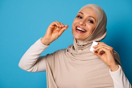 Cute Arab woman with covered head and perfect smile using a dental floss and posing over blue background