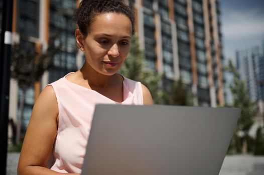 Close-up portrait of middle aged African or Latin American woman, business lady, office manager, worker, employee in casual attire working on laptop on the high buildings background