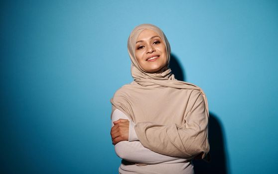 Confident portrait of smiling Arab woman in hijab looking at camera, isolated on blue background. Copy space
