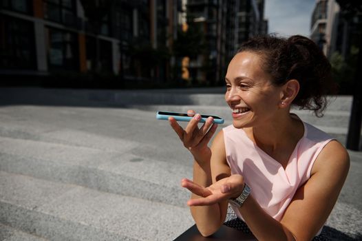 Adorable mixed race beautiful woman recording a voice message on smartphone, resting on steps on the buildings background