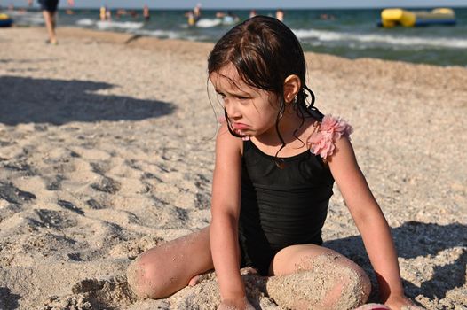 A little cute brunette girl in stylish swimsuit sitting on a sandy beach by the sea and playing with the sand. Child having sunbathing, enjoying beautiful sunny day during summer holidays