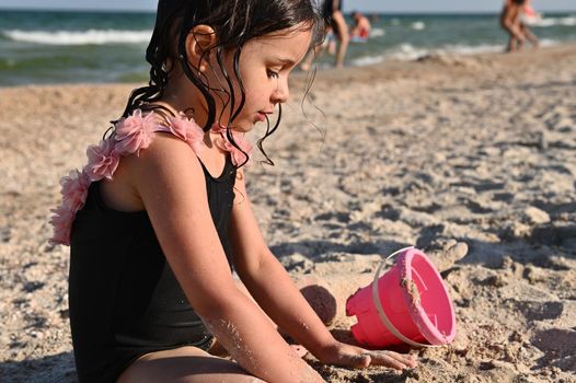 Cute baby girl playing with rake and pink toy bucket with sand, building sandy shapes and castles, enjoying sunbathing and summer vacations. Happy childhood, wellness and recreation concepts