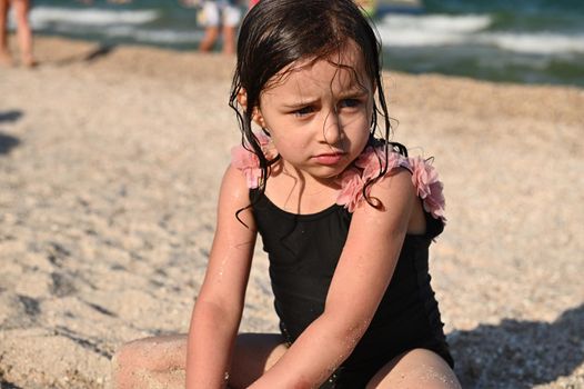 Headshot portrait of cute upset baby girl with wet hair and swimsuit, sitting on the sandy beach on the sea background. Kids emotions. Summer holiday concept