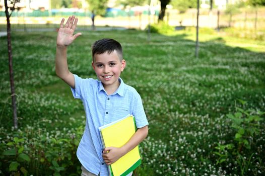 Happy elementary student, pupil, coming back to school. Adorable schoolboy holding a workbooks and waving hello to camera standing on green grass background.