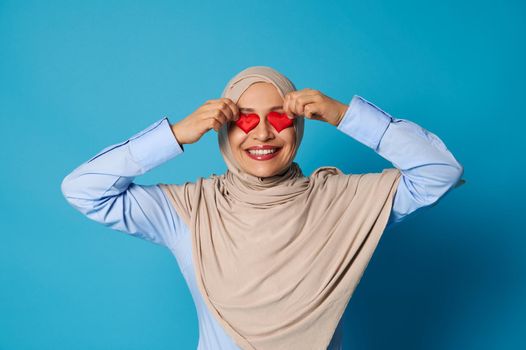 Muslim woman in hijab with oriental appearance putting heart shapes on her eyes and smiling with toothy smile. Copy space