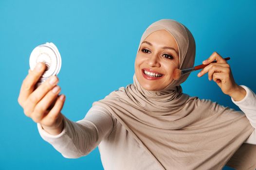 Arab Muslim woman with covered looking at mirror and applying make up, isolated on blue background with copy space