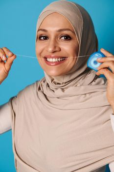 Young Muslim woman with beautiful and healthy smile flossing teeth on blue background with copy space