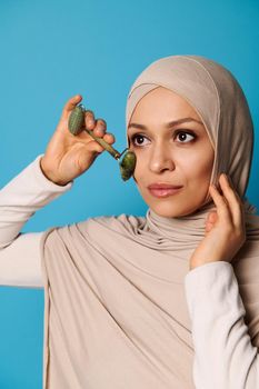 A young beautiful Muslim woman with covered head in hijab doing a facial massage with a jade roller.