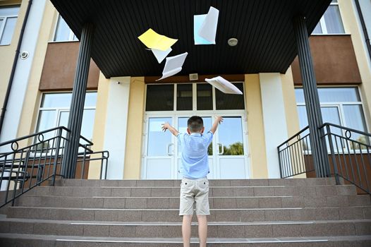 Rear view of schoolboy rejoices at the end of lessons and school, throws workbooks up, laughing and smiling happily while standing on the stairs against the background of the school institution.