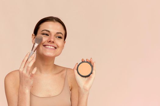 A beautiful woman applies powder using a cosmetic brush on her face skin on a pink background