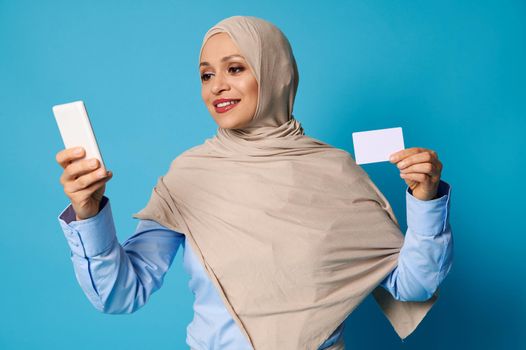 Young woman in hijab holding a white blank plastic card in her hand and texting on mobile phone in her other hand. Isolated on blue background with copy space