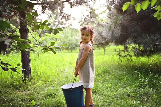 Beautiful little girl in a linen dress stands in the garden with a large blue metal bucket in her hands at sunset