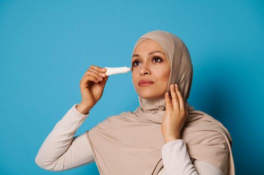 Beauty portrait of Muslim Arab woman with perfect skin applying cream under eyes isolated on blue background with copy space
