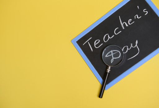 Flat lay composition from a magnifier, magnifying glass, lens, loupe on a chalkboard with lettering Teachers Day on yellow background with copy space for text.