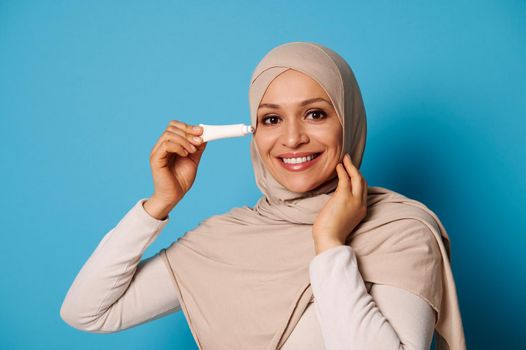 Closeup beauty portrait of woman of oriental appearance applying an under eyes anti aging cream, smiling while posing into camera over blue background with copy space