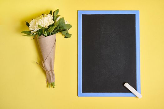 Flat lay composition of delicate beautiful stylish bouquet of asters flowers in craft wrapping paper tied with a rope and blank empty chalkboard with space for text isolated on yellow background