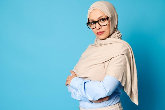 Side portrait of a beautiful Arab Muslim woman with covered head in hijab and wearing eyeglasses isolated over blue background with copy space