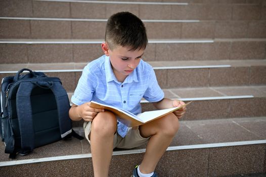 Puzzled elementary school student, pupil, school boy, doing his homework on a notebook while sitting on the stairs of a school institution. Study difficulties, back to school concept