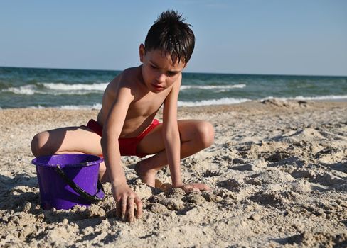 Handsome teenager in red swimming trunks playing on the sandy beach. Adorable male kid builds sand figures with wet sand against the background of the sea. Summer holidays concept. Active lifestyles and happy summer vacations