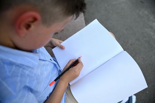 Overhead view of school boy writing on white blank paper sheet of workbook. Copy space. Back to school. Top view