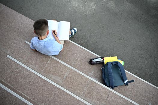 A schoolboy concentrated on doing homework, sitting on the stairs of school building next to his pencil case with school supplies and backpack with workbooks on the foreground. Back to school concept.
