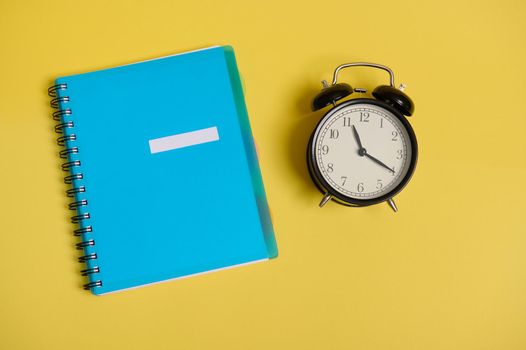 High angle view of a blue colorful notepad, organizer and an alarm clock, on yellow background with space for text. Back to School and Teachers' Day, office and business Concepts. Flat lay