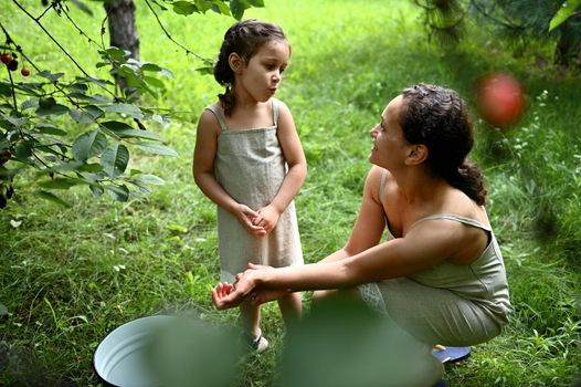 Adorable girl with her charming mother eating cherries while picking in the garden. Happy motherhood and childhood, love, tenderness. Enjoying time together. Happy family. Positive human emotions.