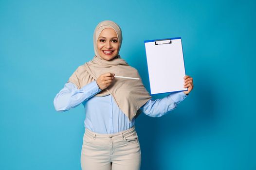 Isolated portrait of a charming Muslim woman in hijab with a beautiful smile points a pen on a white blank sheet of paper on the clipboard