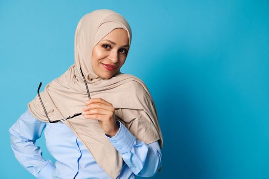 Confident portrait of an attractive Arab woman with covered head in hijab and wearing casual clothes on blue background with copy space